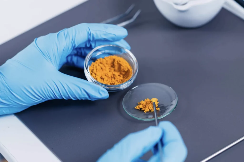 Gloved hands handling a turmeric-coloured powder in laboratory glassware