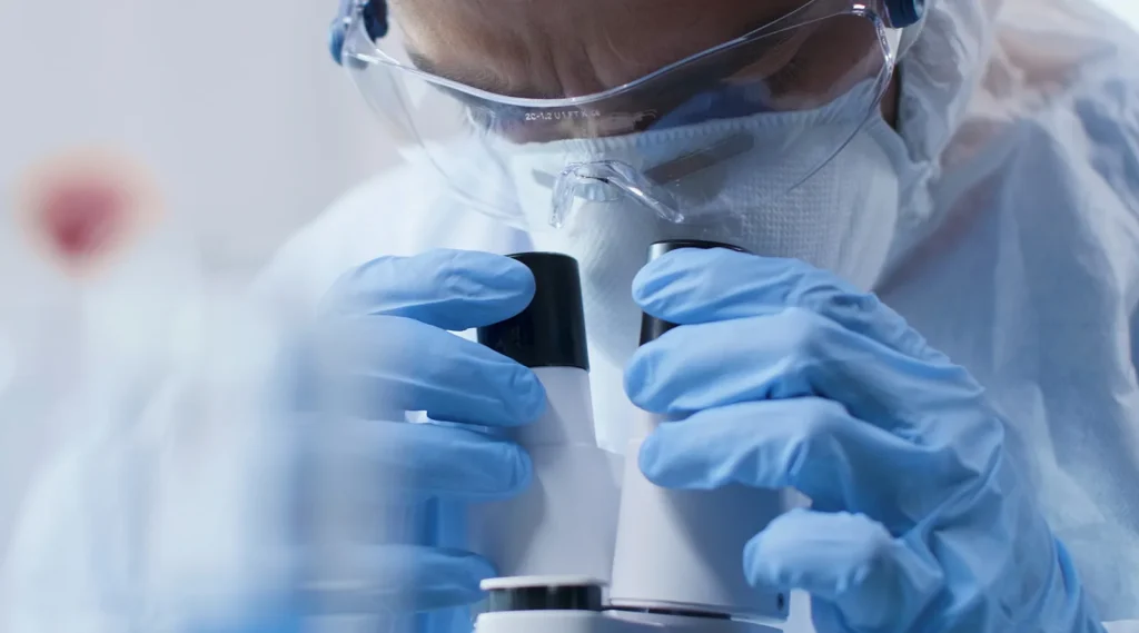 A researcher in protective clothing looking through a microscope